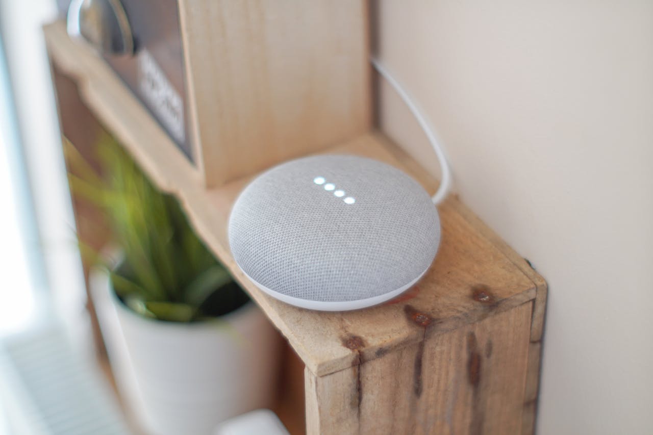 about-img-01 A Google Home Mini smart speaker on a wooden shelf, blending technology with home decor.
